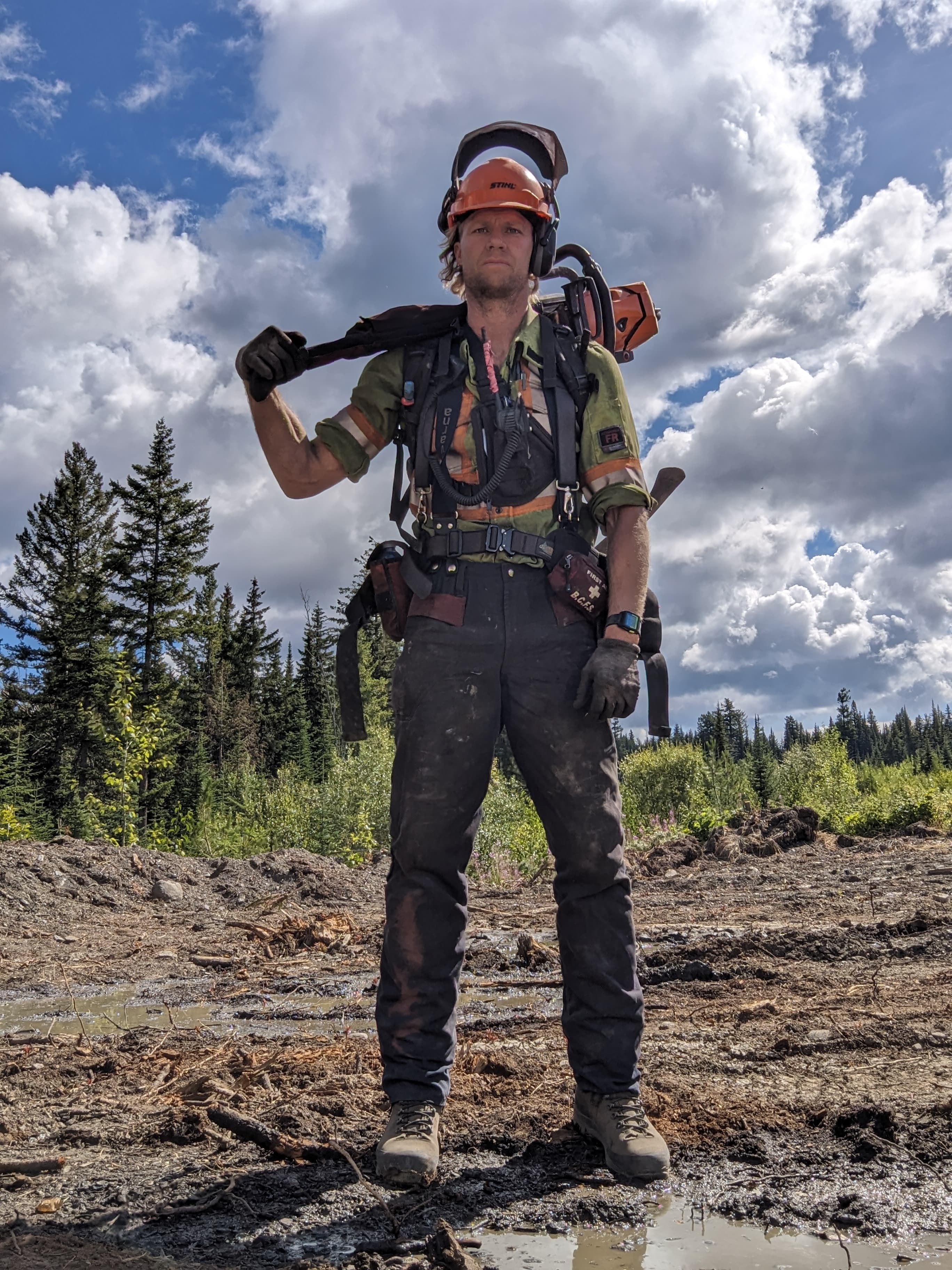 Dirty wildland firefighter in protective gear stands in a muddy forest clearing, carrying a chainsaw.